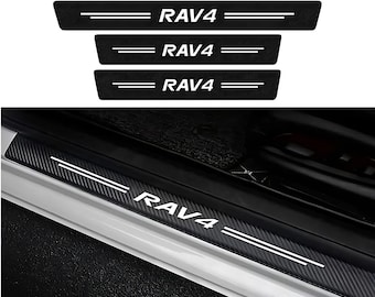 May include: A set of four black door sill protectors with a carbon fiber texture and white RAV4 lettering. Each rectangular strip features two horizontal white lines flanking the text, designed for installation on vehicle door entryways.