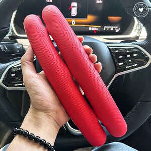May include: Two bright red, perforated car steering wheel covers are held in front of a car's dashboard and steering wheel. The covers are cylindrical and appear to be made of leather or a similar material. The car's interior is visible in the background.