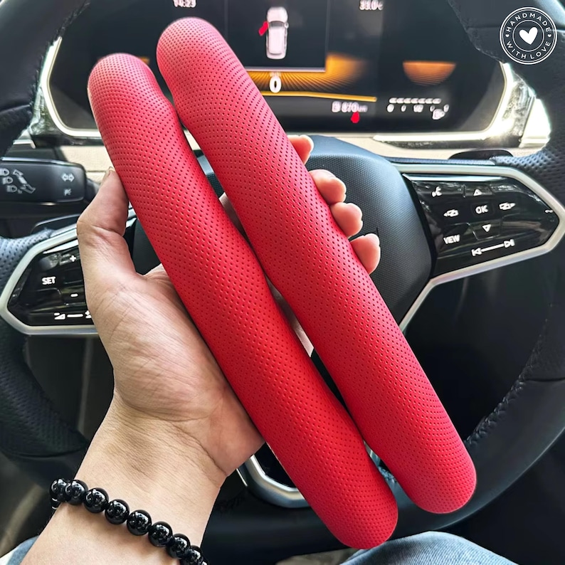 May include: Two bright red, perforated car steering wheel covers are held in front of a car's dashboard and steering wheel. The covers are cylindrical and appear to be made of leather or a similar material. The car's interior is visible in the background.