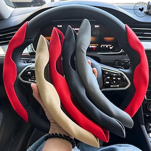 May include: A collection of steering wheel covers in various colors, including beige, red, black, and gray. The covers are held in front of a car's steering wheel, which has a red and black design. The image is well-lit, showcasing the textures and colors of the covers.