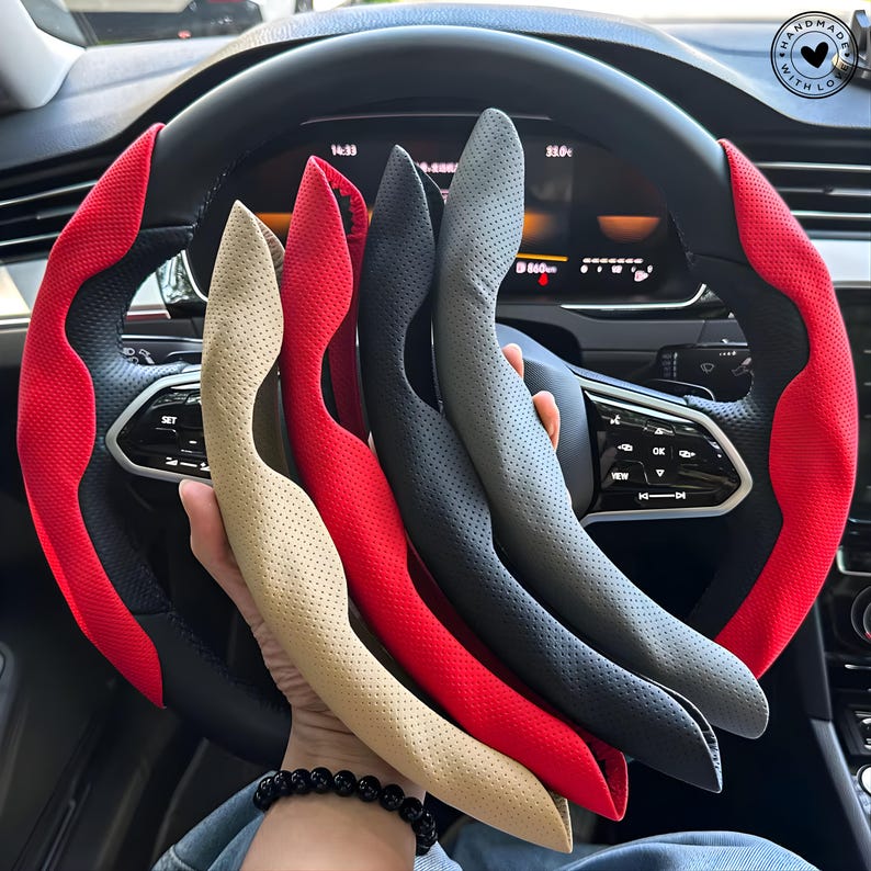 May include: A collection of steering wheel covers in various colors, including beige, red, black, and gray. The covers are held in front of a car's steering wheel, which has a red and black design. The image is well-lit, showcasing the textures and colors of the covers.