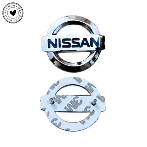 Peut inclure: Un emblème Nissan chromé avec le nom de la marque en lettres bleues. L'emblème est circulaire avec une barre rectangulaire au centre. En dessous se trouve le dos de l'emblème avec un adhésif 3M.