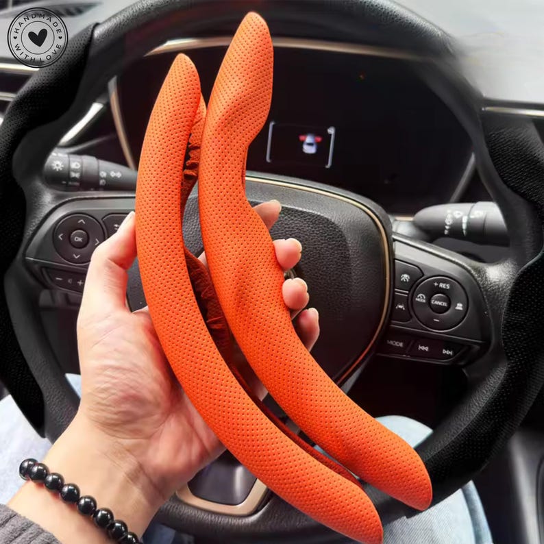 May include: Two orange steering wheel covers with a perforated texture are held in front of a car's steering wheel. The covers are curved and designed to fit the steering wheel's shape. The car's dashboard and steering wheel are visible in the background.