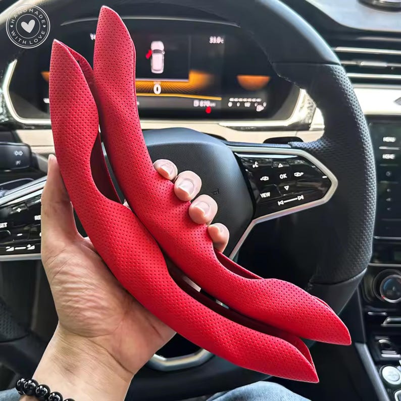 May include: Two bright red, perforated car steering wheel covers. The covers are held up in front of a car's steering wheel, revealing the interior dashboard and controls. The covers are made of a soft, textured material.