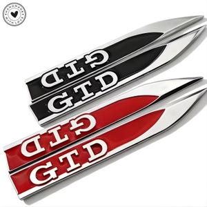 Könnte beinhalten: Zwei Paare von GTD-Autoemblemen. Ein Paar ist schwarz mit silberner Umrandung und weißen Buchstaben, das andere ist rot mit silberner Umrandung und weißen Buchstaben. Die Embleme sind länglich und haben ein spitzes Ende.