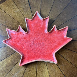 Peut inclure: Un plat en céramique rouge en forme de feuille d'érable avec une surface texturée. Le plat a un bord légèrement surélevé et est posé sur un fond en bois. Cette pièce décorative est idéale pour servir des collations ou comme élément décoratif.