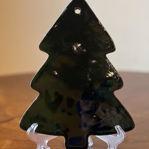 May include: A glossy black Christmas tree ornament. The tree-shaped decoration has a hole at the top for hanging. It is displayed on a clear plastic stand, showcasing its reflective surface and festive design.