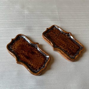 Puede incluir: Dos reposacucharas de cerámica marrón con un borde decorativo festoneado. Los reposacucharas tienen un acabado brillante y moteado con un borde marrón más oscuro. El fondo es una tela beige claro.