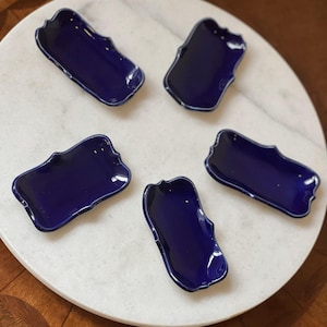 Peut inclure: Cinq petits plats en céramique bleu marine avec un design de bordure festonné unique. Les plats sont disposés sur une surface en marbre blanc, mettant en valeur leur finition brillante. L'esthétique générale est moderne et élégante.