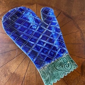Puede incluir: Un guante de cocina de cerámica en un tono azul vibrante con un patrón de diamantes. El borde inferior presenta un diseño verde y ondulado. El guante está colocado sobre una superficie de madera.