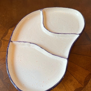 Puede incluir: Una fuente de cerámica blanca con tres secciones y un borde morado. La fuente tiene un diseño moteado con pequeños puntos de varios colores. Las secciones están divididas por líneas curvas, creando una forma única.