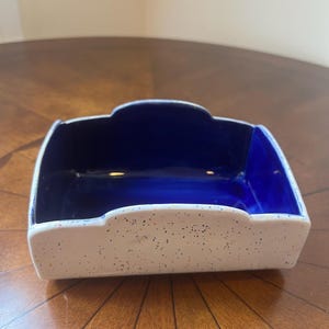 May include: A rectangular ceramic napkin holder with a white speckled exterior and a deep blue interior. The holder has a scalloped edge design and is placed on a wooden surface.