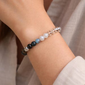 Peut inclure: Un bracelet de perles avec un mélange de pierres précieuses colorées et de perles transparentes. Le bracelet comprend des perles bleues, blanches et transparentes. Le mot "KOSMIK" est imprimé en rouge en bas de l'image.