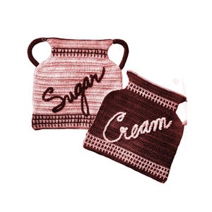 May include: Two crocheted cream and sugar containers. The cream container is brown with a pink cream label. The sugar container is pink with a brown sugar label.
