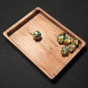 May include: A wooden dice tray with a rectangular shape and raised edges. Several polyhedral dice with green and gold coloring are scattered inside the tray. The dice have white numbers. The tray is made of light brown wood.