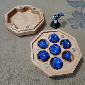 May include: A wooden dice box with a set of blue and gold dice. The box is octagonal and has a magnetic closure. A miniature figurine stands nearby. The dice are a deep blue with gold numbering.