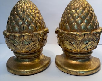 Puede incluir: Un par de remates decorativos de color dorado con forma de piña. Cada pieza presenta una parte superior texturizada similar a escamas que descansa sobre una base adornada con patrones de hojas talladas, con un acabado de pintura dorada.