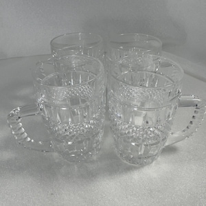 May include: Four clear glass beer mugs with textured diamond patterns and handles. The mugs are arranged in a group, showcasing their design and clarity. The glassware is suitable for serving beverages.