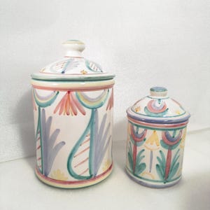 Puede incluir: Dos botes de cerámica con tapas. El bote más grande es cilíndrico y el más pequeño ovalado. Ambos son blancos con diseños florales y geométricos coloridos en rosa, azul, amarillo y verde. Las tapas tienen pequeños pomos.