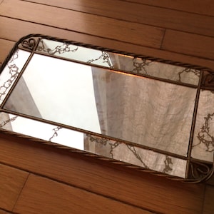 May include: A gold-toned rectangular mirror with a decorative, rope-like frame. The mirror is divided into four sections with a pattern of small, intricate designs.