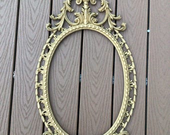 May include: A gold oval-shaped mirror frame with ornate details. The frame has a decorative top with a scroll design and a beaded border.