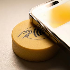 Può includere: Uno smartphone è appoggiato su un dispositivo di pagamento NFC giallo. Il dispositivo ha un simbolo di mano e onde. Lo schermo del telefono mostra una sfumatura gialla. Lo sfondo è beige chiaro.