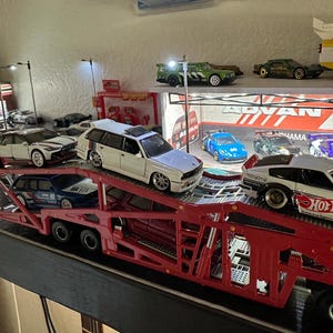 May include: A red car hauler carrying several toy cars, including a white station wagon and a red and white sports car. Other toy cars are on a shelf in the background. The scene is well-lit.
