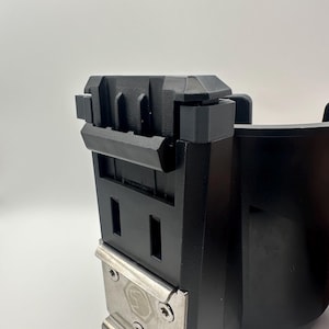 May include: Close-up of a black plastic object with a rectangular shape and a metal plate attached. The metal plate is secured with screws and features a circular emblem. The object has a series of rectangular openings.