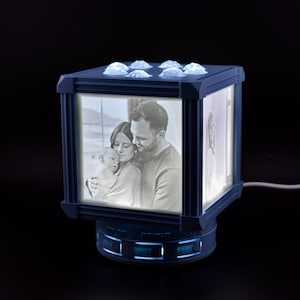 May include: A navy blue photo cube lamp with illuminated family photos. The lamp has a square shape with a base that reads "COLOR BRIGHT SUN". The top of the lamp has several clear, rounded accents. A white cord extends from the base.