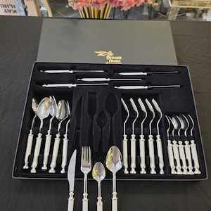 May include: A complete silver-toned flatware set with white handles, neatly arranged in a black box. The set includes knives, forks, spoons, and serving utensils. The box is labeled "Dream Home" in gold lettering. A vase of pink flowers is in the background.
