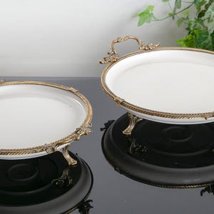 以下が含まれることがあります： 金色の縁取りと装飾的な取っ手がついた白いセラミック製のサービングトレイが2つ。トレイは3本の金色の脚で支えられており、黒い表面に置かれています。