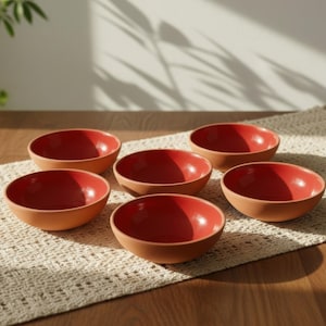 May include: Six terracotta-colored bowls with a vibrant red interior are arranged on a cream-colored woven placemat. The bowls are evenly spaced on a wooden surface, with natural light casting shadows.
