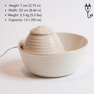 May include: Cream ceramic pet fountain with a tiered, conical water feature and a wide, ribbed bowl. The fountain is 7 cm high, 22 cm wide, and weighs 2.5 kg. It has a capacity of 1.5 L. A power cord extends from the fountain.