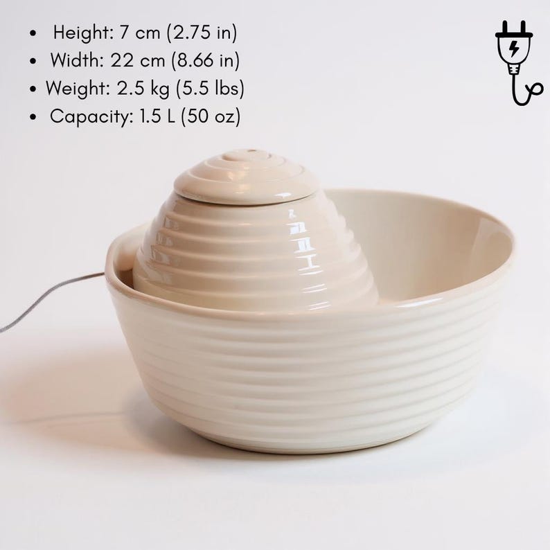 May include: Cream ceramic pet fountain with a tiered, conical water feature and a wide, ribbed bowl. The fountain is 7 cm high, 22 cm wide, and weighs 2.5 kg. It has a capacity of 1.5 L. A power cord extends from the fountain.