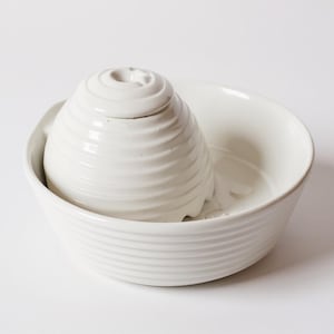 May include: A white ceramic pet water fountain. It features a spiral-shaped top and a bowl-shaped base. The top has a small star detail. The fountain is designed to provide fresh water for pets.