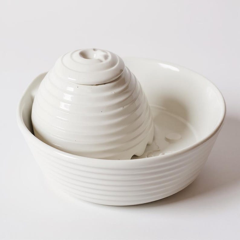 May include: A white ceramic pet water fountain. It features a spiral-shaped top and a bowl-shaped base. The top has a small star detail. The fountain is designed to provide fresh water for pets.
