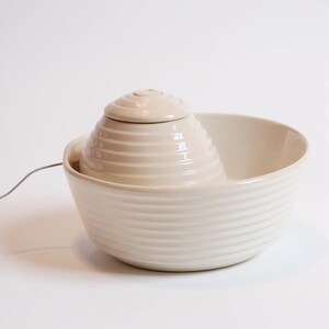 May include: A cream ceramic pet water fountain. The fountain has a tiered, cone-shaped centre piece with a small lid, set within a larger, round bowl. The bowl and centre piece have a ribbed design.