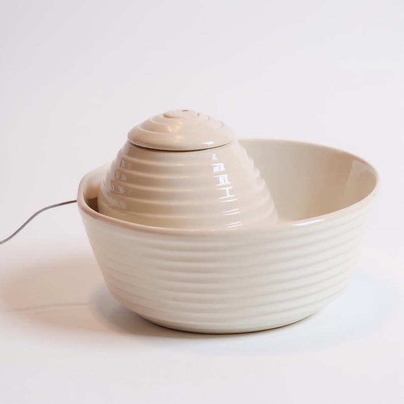 May include: A cream ceramic pet water fountain. The fountain has a tiered, cone-shaped centre piece with a small lid, set within a larger, round bowl. The bowl and centre piece have a ribbed design.