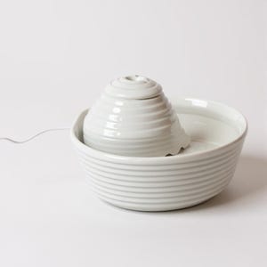 May include: A white ceramic pet water fountain. It features a tiered, cone-shaped fountain head and a wide, shallow bowl. The fountain head has a small opening at the top. The bowl and fountain head have horizontal ridged detailing. A white cord extends.