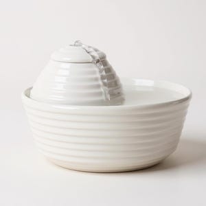 May include: A white ceramic pet water fountain. The fountain has a tiered design, with a central, cone-shaped element from which water flows into a wide, ribbed bowl. The water is clear and the fountain is set against a white background.