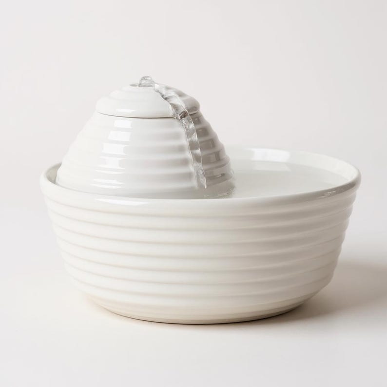 May include: A white ceramic pet water fountain. The fountain has a tiered design, with a central, cone-shaped element from which water flows into a wide, ribbed bowl. The water is clear and the fountain is set against a white background.