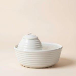 May include: A white ceramic pet fountain with a tiered, ridged design. The fountain has a water basin and a central, cone-shaped feature with a small spout. The overall aesthetic is minimalist and modern.