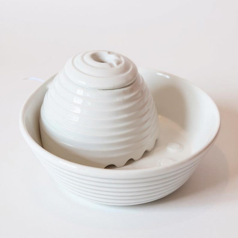 May include: A white ceramic pet water fountain. It features a tiered, beehive-shaped central element and a surrounding bowl. The fountain has a minimalist design with a ridged texture on the central piece and bowl.