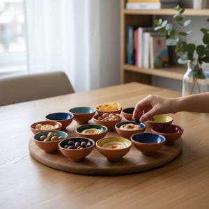 May include: A wooden serving platter displays several small terracotta bowls, each containing different snacks. The bowls have coloured rims, including blue, green, and yellow. The snacks include olives, hummus, and crackers.