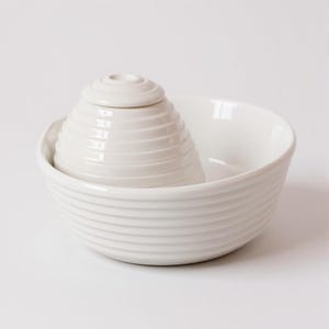 May include: A white ceramic bowl with a ridged exterior contains a smaller, ridged, white ceramic container with a lid. The bowl and container are nested. The background is plain white.