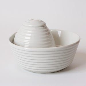 May include: A white ceramic citrus juicer set. The set includes a bowl with horizontal ridges and a conical reamer with a ridged exterior. The juicer is designed for easy citrus fruit juicing.