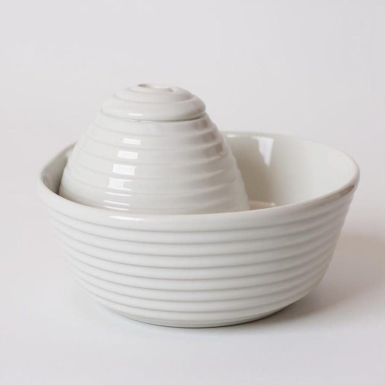 May include: A white ceramic citrus juicer set. The set includes a bowl with horizontal ridges and a conical reamer with a ridged exterior. The juicer is designed for easy citrus fruit juicing.