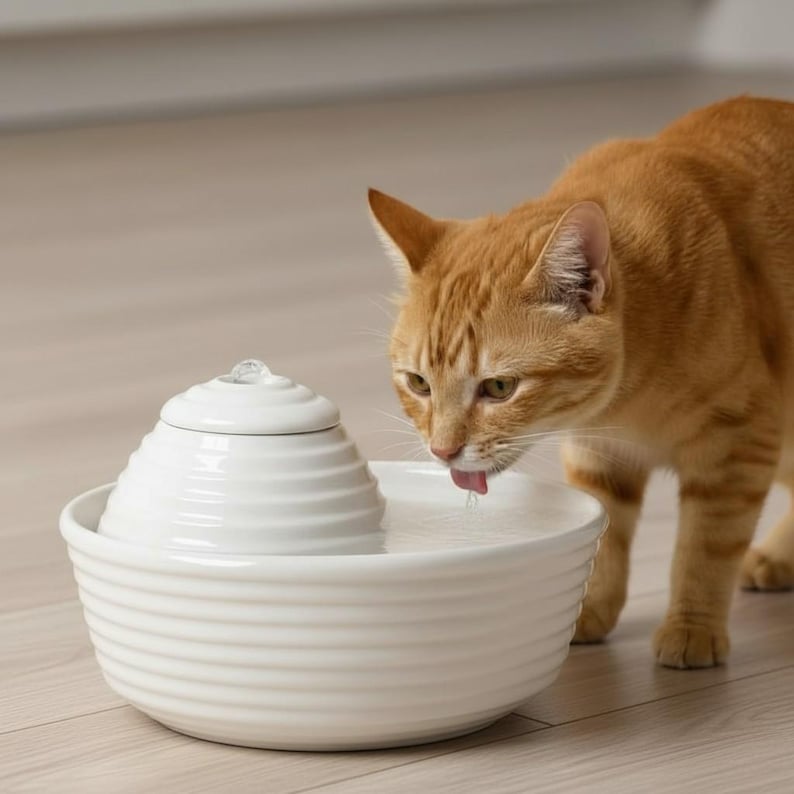 May include: A white ceramic cat water fountain with a tiered design, next to an ginger tabby cat drinking water. The fountain has a round base and a small, decorative top. The cat is in focus, with its tongue out.