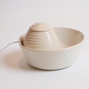 May include: A cream-coloured ceramic water fountain with a tiered, conical top and a wide, shallow bowl. The fountain has a ridged texture and a small spout for water flow. A thin, grey cord extends from the fountain.