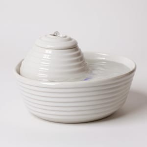 May include: A white ceramic pet fountain with a tiered, spiral design. The fountain has a bowl-shaped base and a central, cone-shaped feature with water gently flowing from the top. The water is clear, and the overall aesthetic is clean and modern.